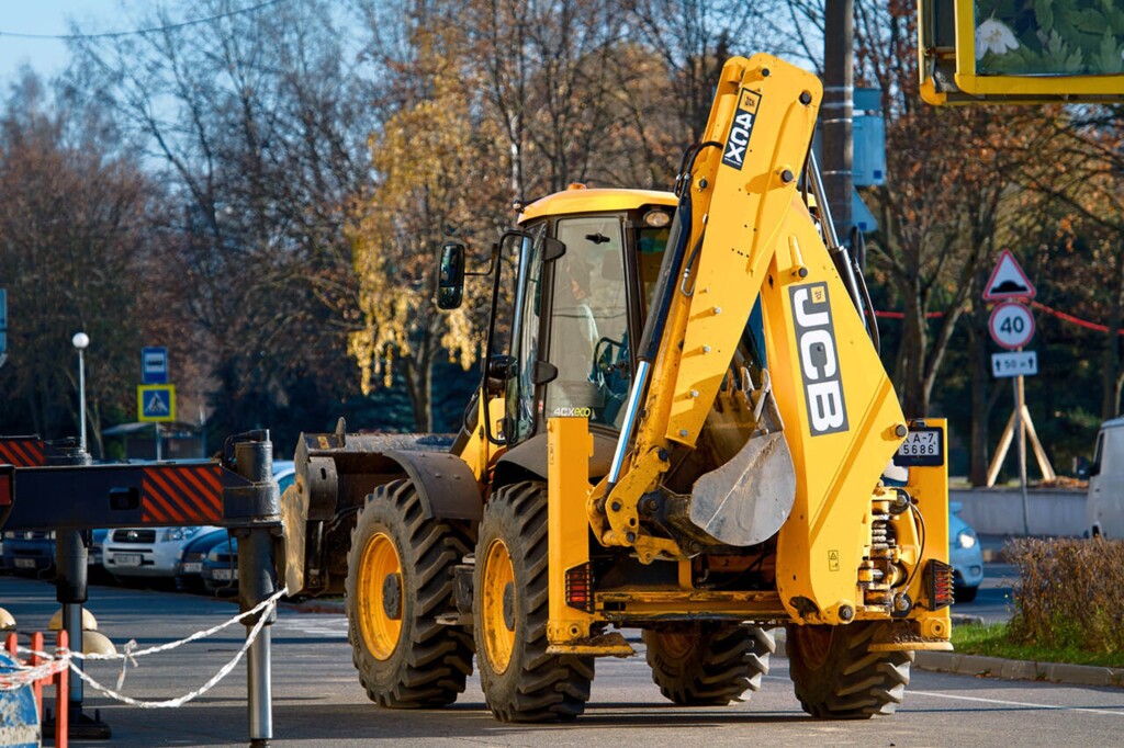 Жовтий екскаватор-навантажувач JCB 4CX працює на будівельному майданчику біля вулиці, в оточенні дерев і дорожніх знаків.