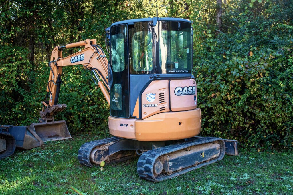 На фото зображено невеликий гусеничний екскаватор CASE, модель CX36B, припаркований на траві поруч з густим чагарником.