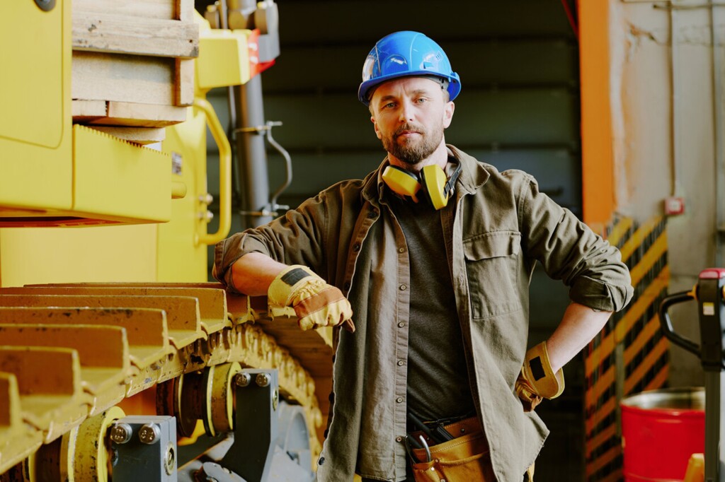 Будівельний робітник у синьому захисному шоломі та робочих рукавичках, спирається на жовте шасі будівельної машини в майстерні.