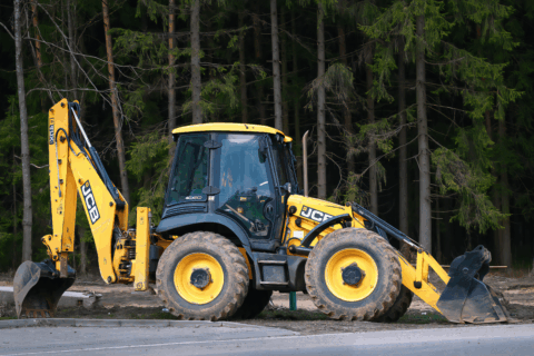 Екскаватор JCB 4CX ECO стоїть на узбіччі дороги в оточенні дерев.
